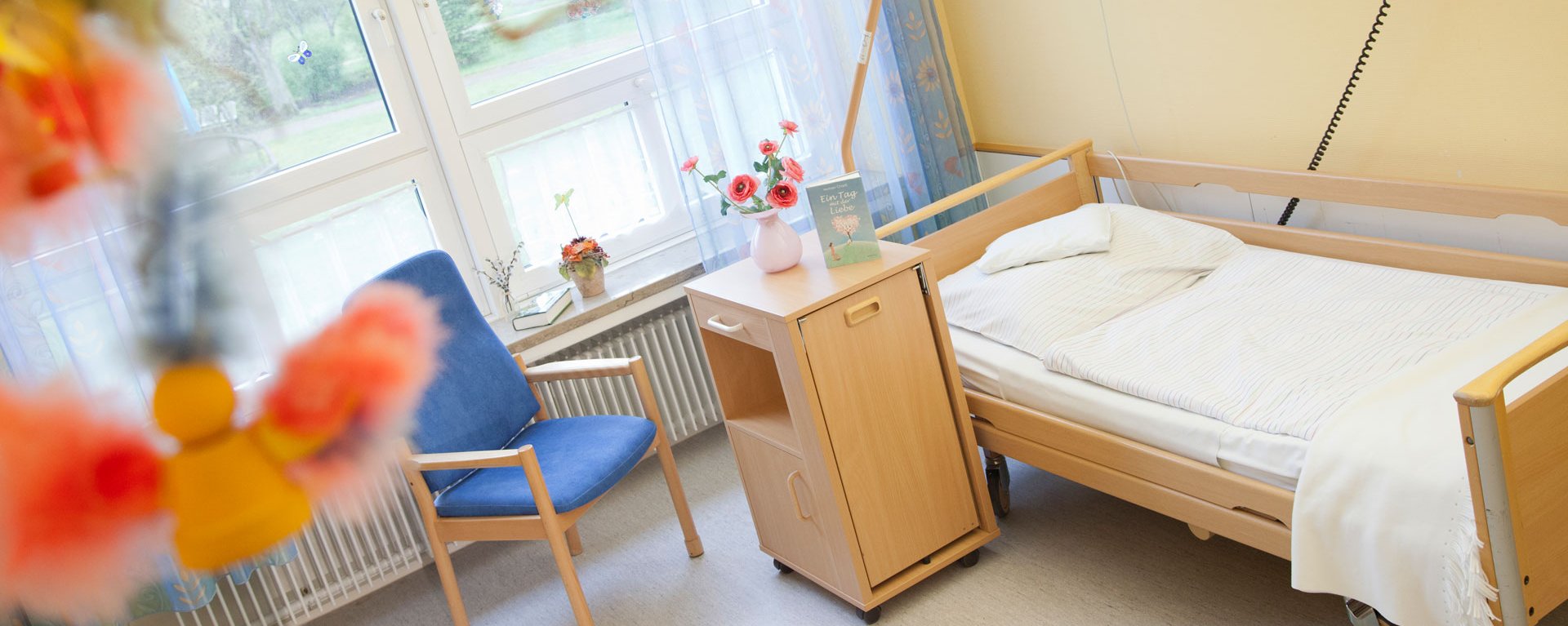 Aufnahme eines Zimmers der modernen Kurzzeitpflege an der Klinik Mindelheim mit Pflegebett, Besucherstuhl großen Fenstern und bunten Blumen unscharf im Vordergund