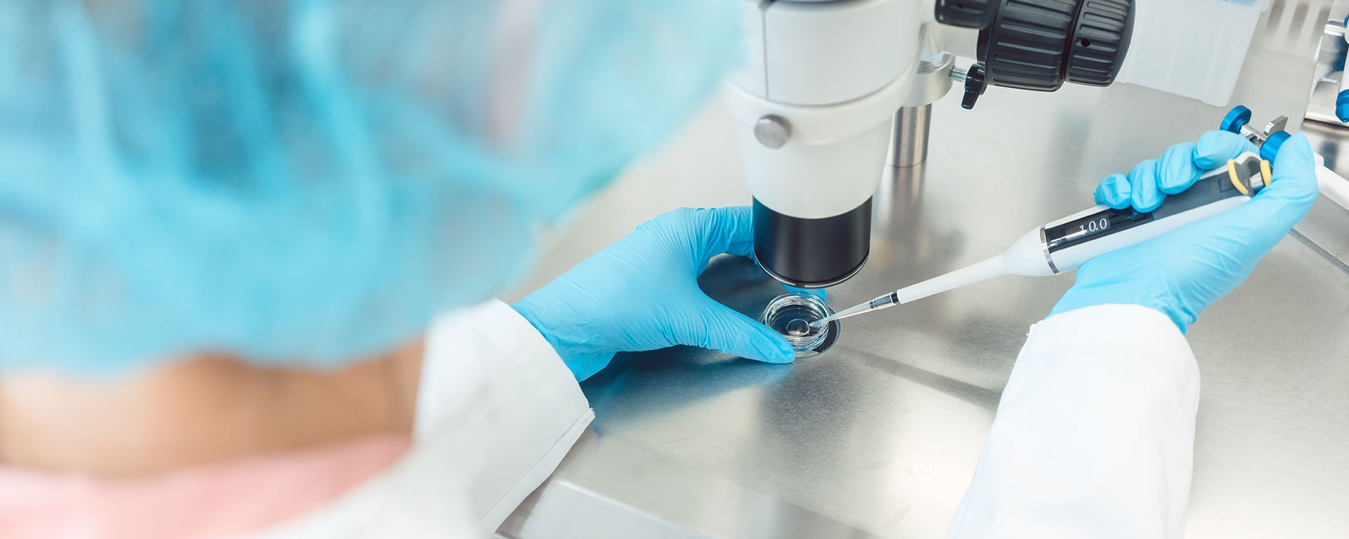 Eine Person, mit Haarhaube, weißem Kittel und Einweghandschuhen, arbeitet mit einer Pipette an einem Mikroskop.