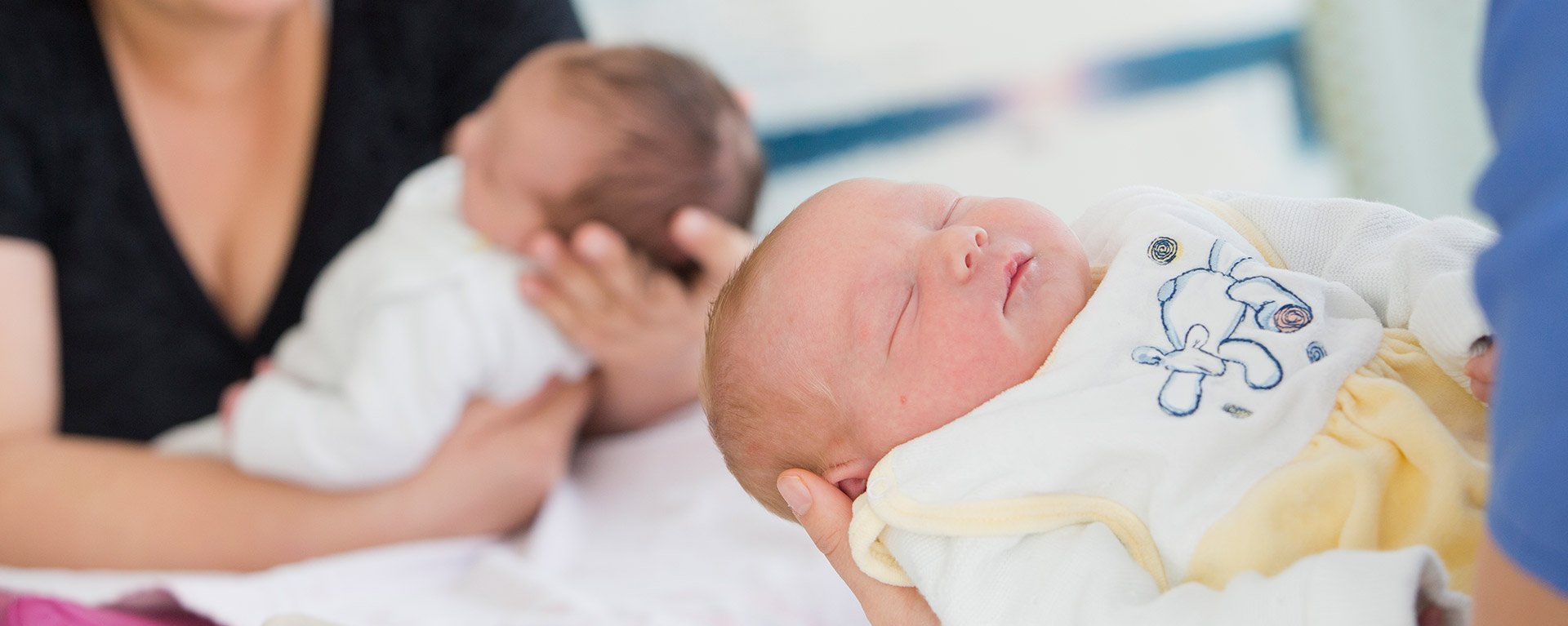 Rechts vorne hält eine Geburtshelferin ein Baby in der Hand. Links im Hintergrund hält eine Mutter ihr Baby in der Hand in der Geburtshilfe der Klinik Mindelheim.